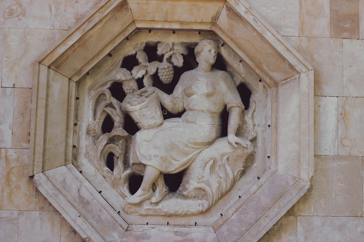 Sculpture en relief représentant une figure féminine assise tenant une corne d'abondance avec fruits et végétation, intégrée dans un médaillon hexagonal en pierre claire sur façade architecturale